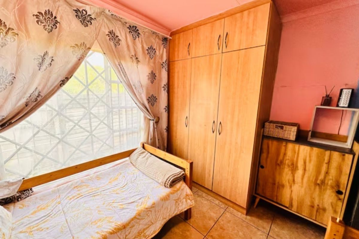 A bedroom featuring built-in wooden wardrobes, tiled flooring, and a window with security bars. The room requires cosmetic updates to the wall finishes.