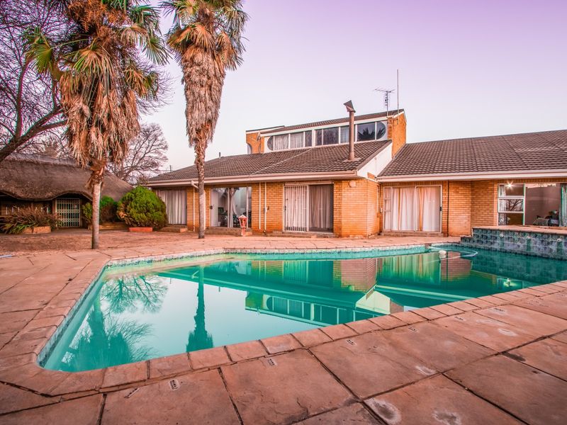 Rear exterior view featuring a swimming pool with tiled surround, brick facade, and a secondary thatched roof structure in the garden.