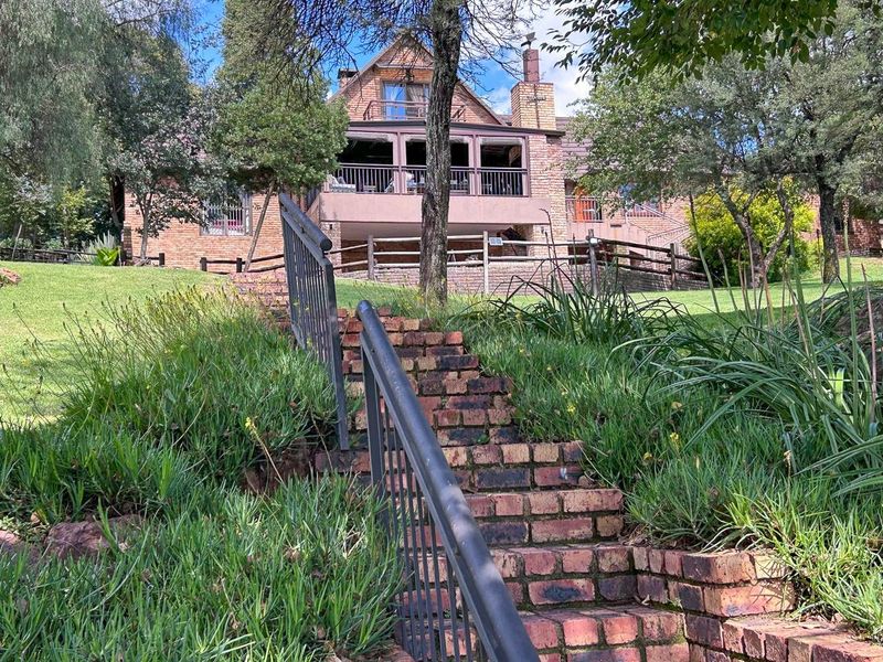 Rear view of a multi-story brick house featuring a covered balcony, chimney, and a brick staircase leading through the landscaped garden.
