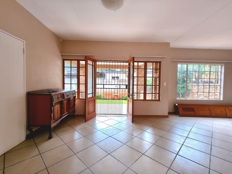 Spacious living area featuring tiled flooring, large wooden-framed windows, and direct access to the garden through a secure double door entrance.