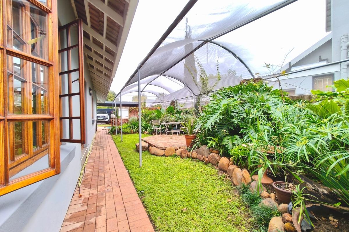 A well-maintained rear garden featuring a brick pathway, stone-edged garden beds, and a large overhead shade cloth structure extending from the house.
