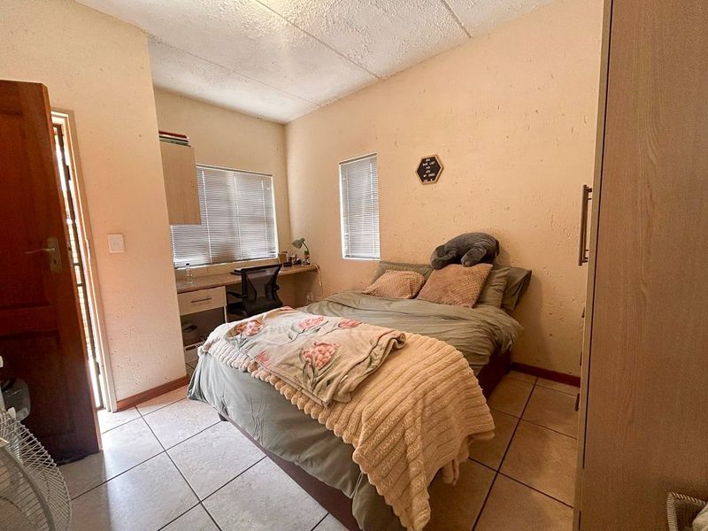 A compact bedroom featuring tiled flooring, built-in desk space, and integrated cupboard storage, with natural light provided by two windows.