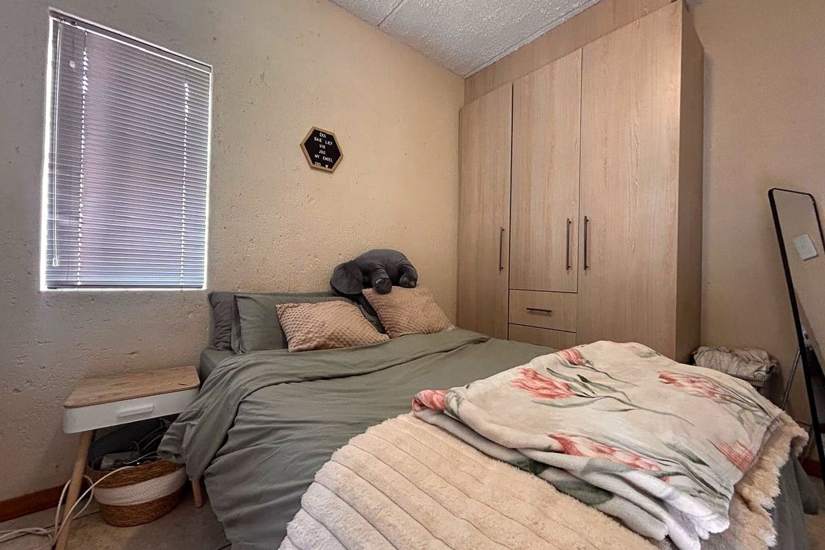 A compact bedroom featuring built-in wooden wardrobes, tiled flooring, and a window fitted with venetian blinds.