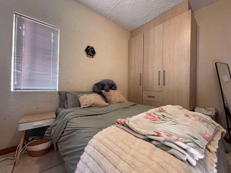 A compact bedroom featuring built-in wooden wardrobes, tiled flooring, and a window fitted with venetian blinds.