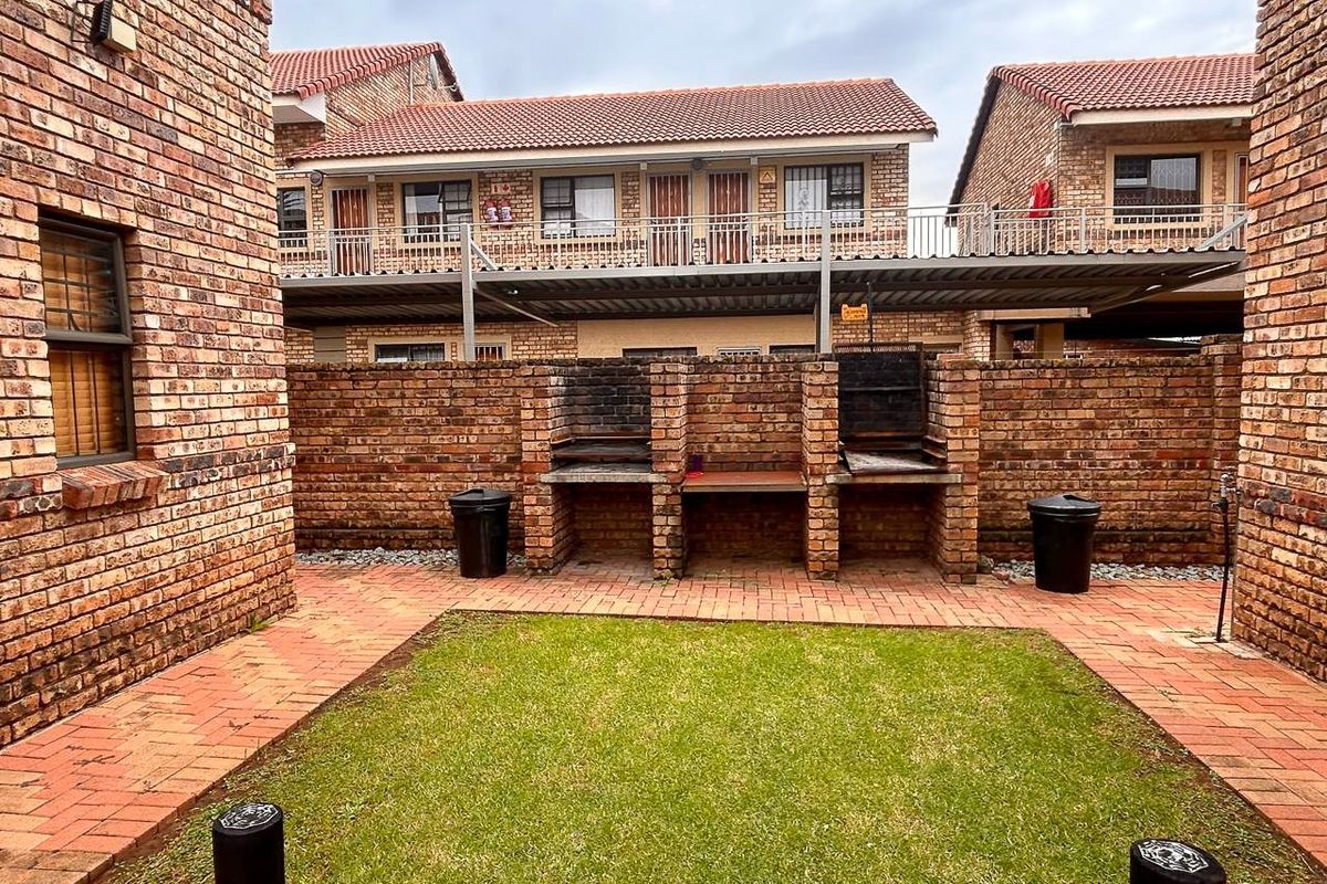 A rear courtyard area featuring a brick-built double braai station, paved flooring, and a small lawn, set against a multi-story apartment building.
