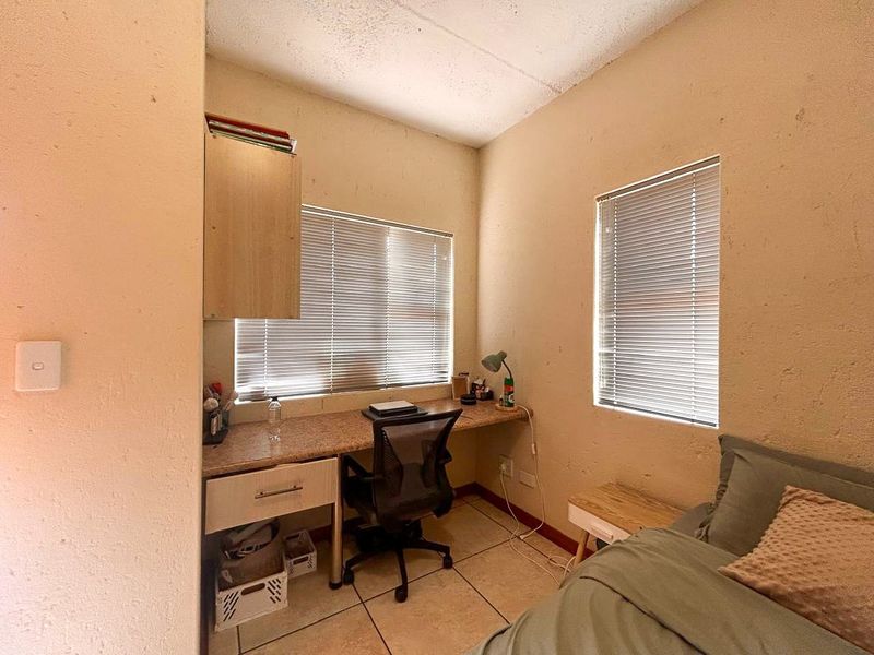 A compact bedroom featuring a built-in desk, integrated cabinetry, tiled flooring, and dual windows with venetian blinds.