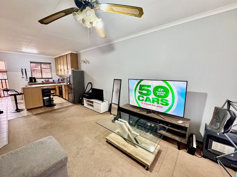 Open-plan living area featuring a carpeted lounge space adjacent to a functional kitchen with built-in cabinetry and tiled flooring.