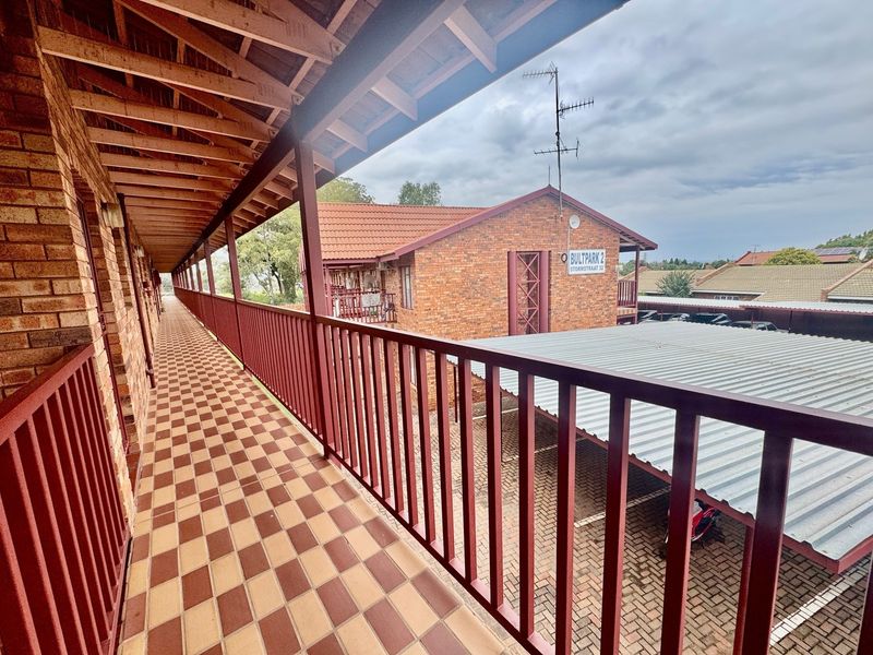 An exterior view of a covered apartment balcony featuring brick walls, tiled flooring, and metal railings overlooking the complex parking area.