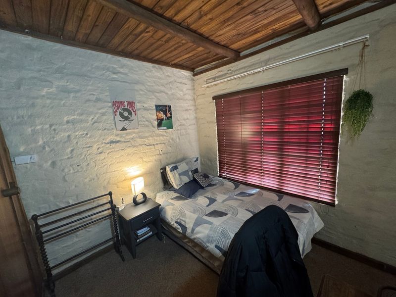 A cozy bedroom featuring rustic painted brick walls and a warm timber beam ceiling, complemented by fitted venetian blinds.