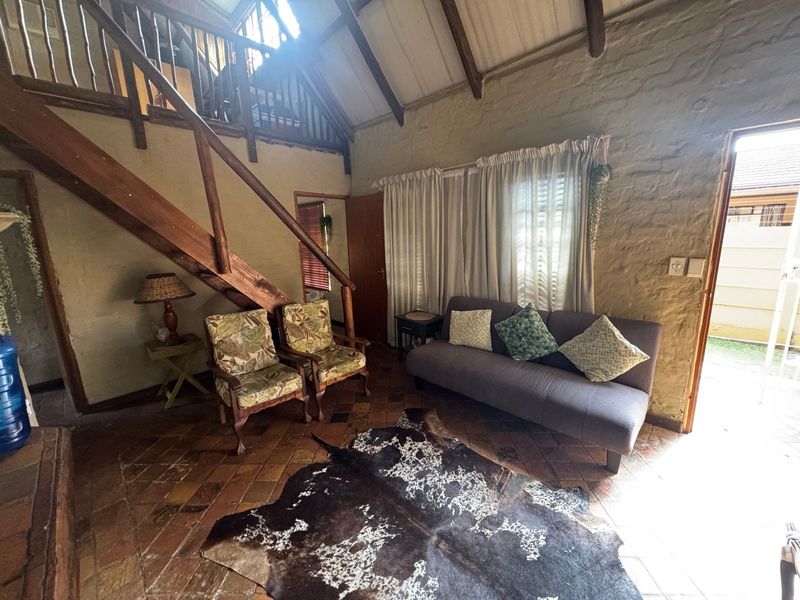 Rustic living room featuring exposed timber beams, a wooden staircase leading to a mezzanine level, and traditional terracotta floor tiles.