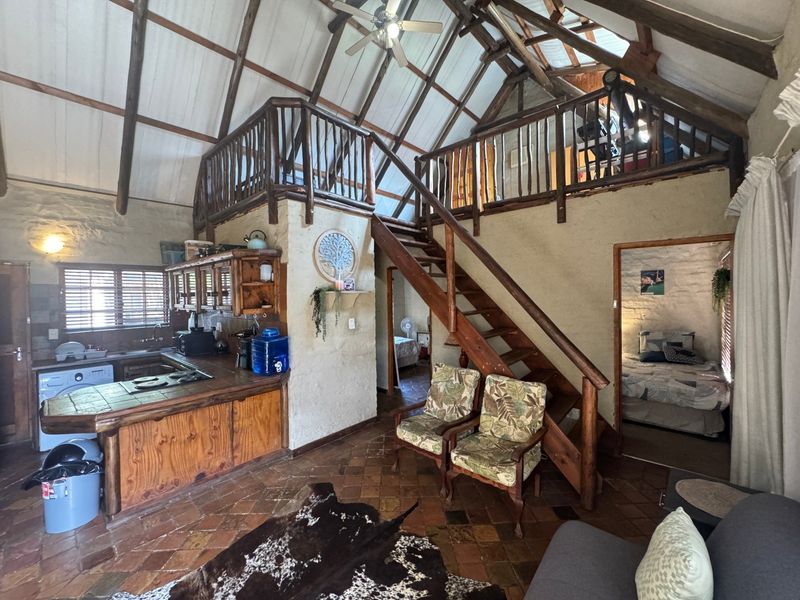 Open-plan living area featuring a rustic kitchen, terracotta flooring, exposed timber rafters, and a mezzanine level accessed by a wooden staircase.