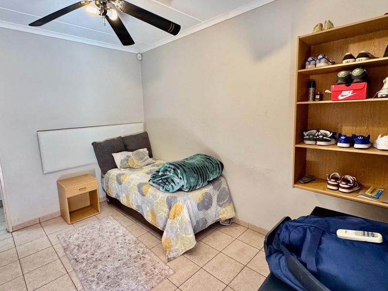 A bedroom featuring tiled flooring, a ceiling fan, and built-in wall shelving, with an adjacent view into a kitchen area.