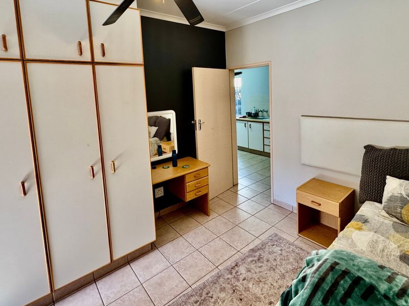 A bedroom featuring built-in cupboards, tiled flooring, and a ceiling fan, with an adjacent view into a kitchen area.