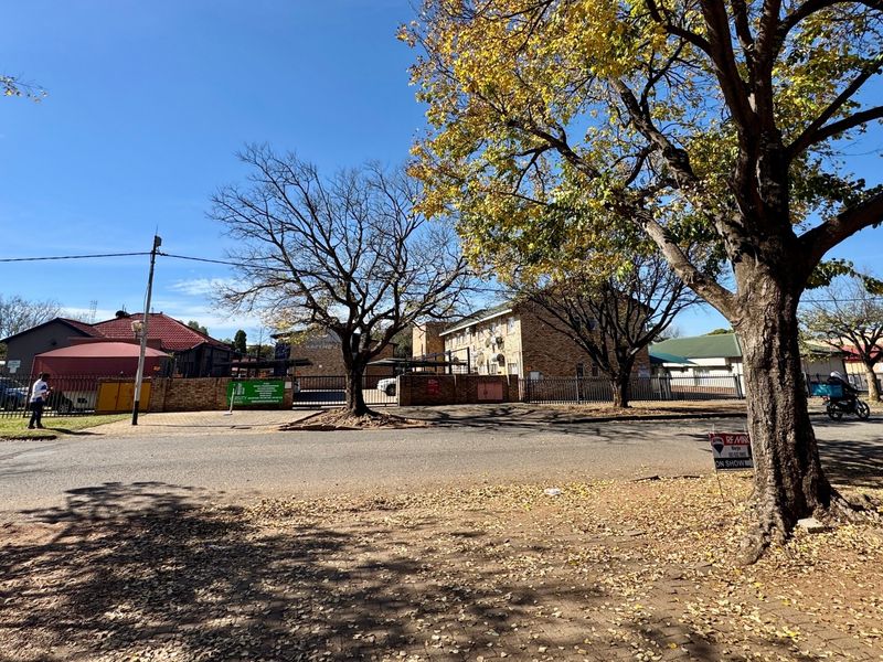 Street view of the apartment complex entrance, featuring mature trees, perimeter fencing, and brick building facades.