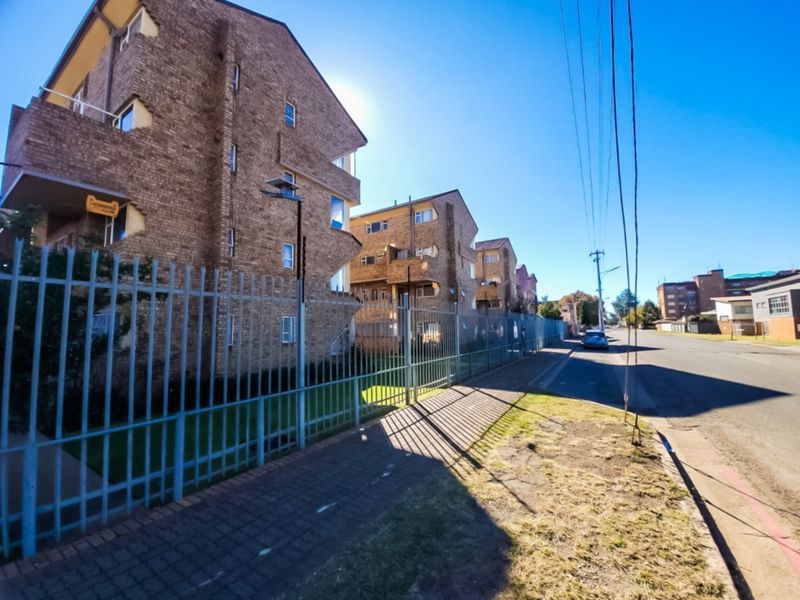 Street-level view of a multi-story brick apartment complex featuring secure metal perimeter fencing and adjacent sidewalk access.