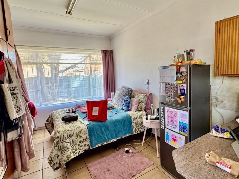 Compact open-plan living space featuring tiled flooring, built-in storage cupboards, and a functional kitchenette area with window security bars.