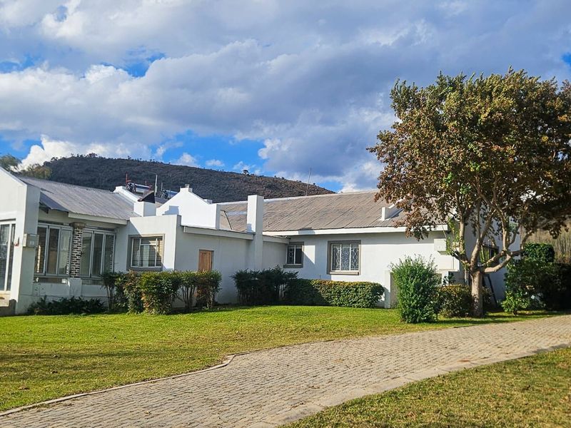 Front view of a single-story small holding residence featuring a paved driveway, corrugated metal roof, and established garden surroundings with mountain views.