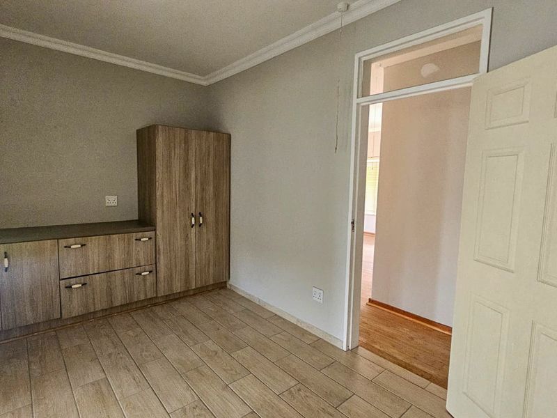 A clean, empty bedroom featuring neutral tiled flooring, built-in wooden cabinetry, and a classic door frame leading to an adjacent hallway.