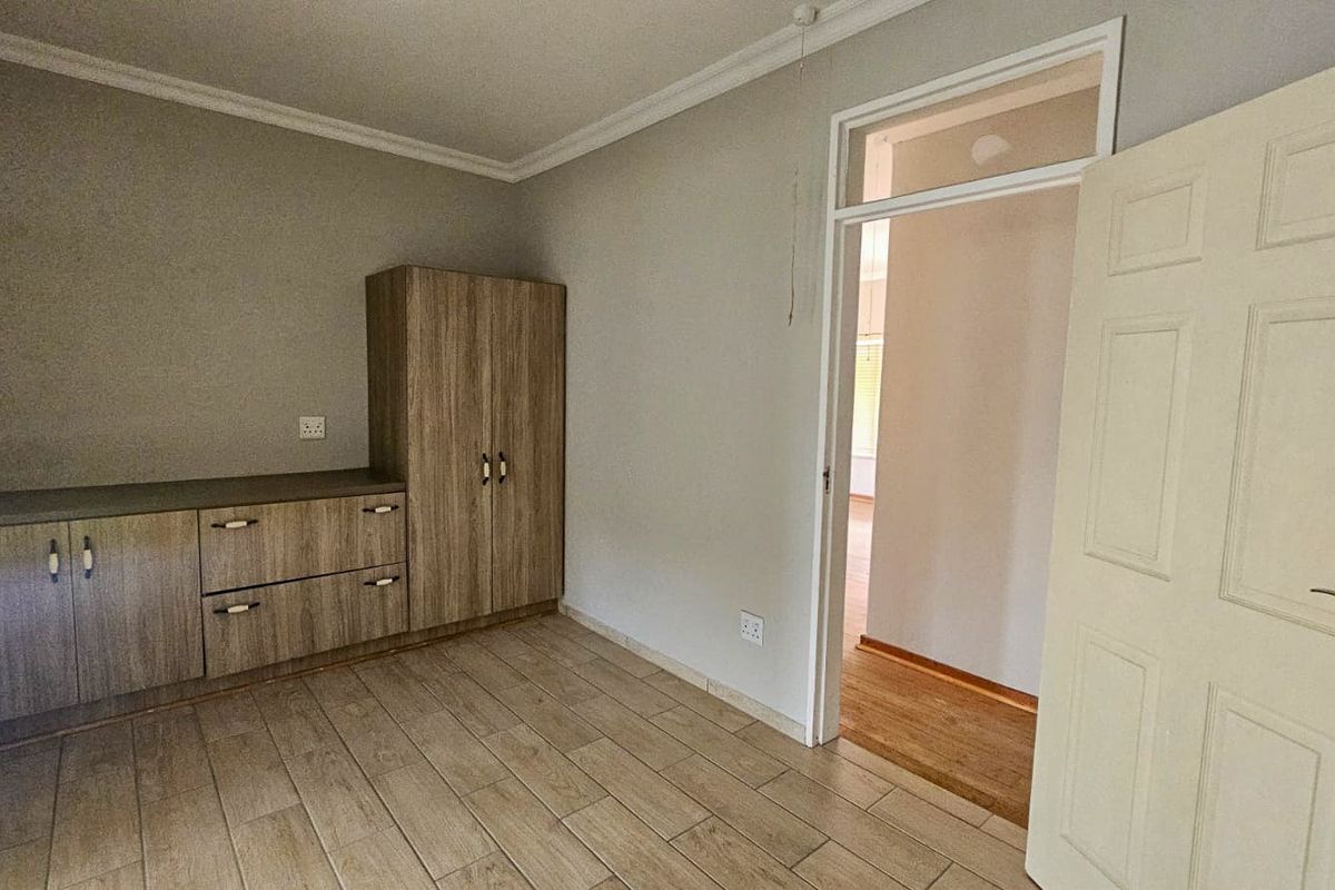 A clean, empty bedroom featuring neutral tiled flooring, built-in wooden cabinetry, and a classic door frame leading to an adjacent hallway.