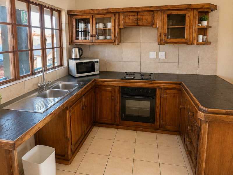A functional kitchen featuring rustic wooden cabinetry, dark tiled countertops, built-in oven and hob, and large timber-framed windows providing natural light.