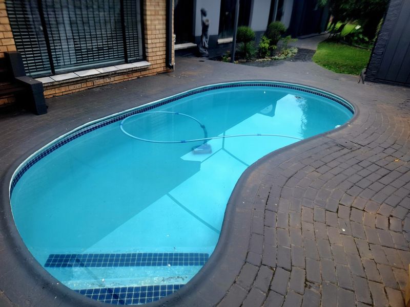 A kidney-shaped swimming pool with blue tiled trim, surrounded by dark brick paving and adjacent to the main house structure.