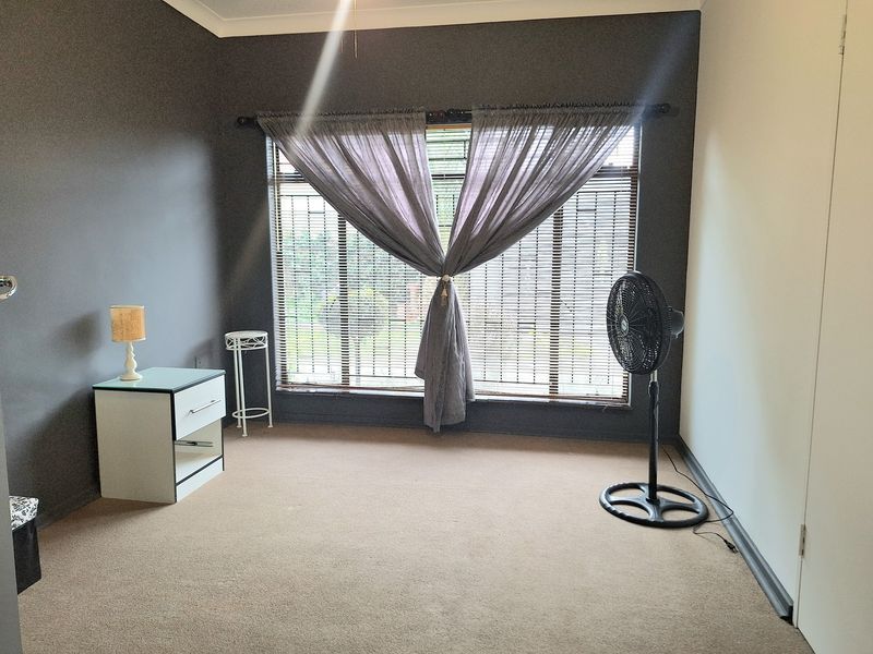 A spacious bedroom featuring neutral carpeting, dark accent walls, and large windows fitted with venetian blinds and built-in storage.