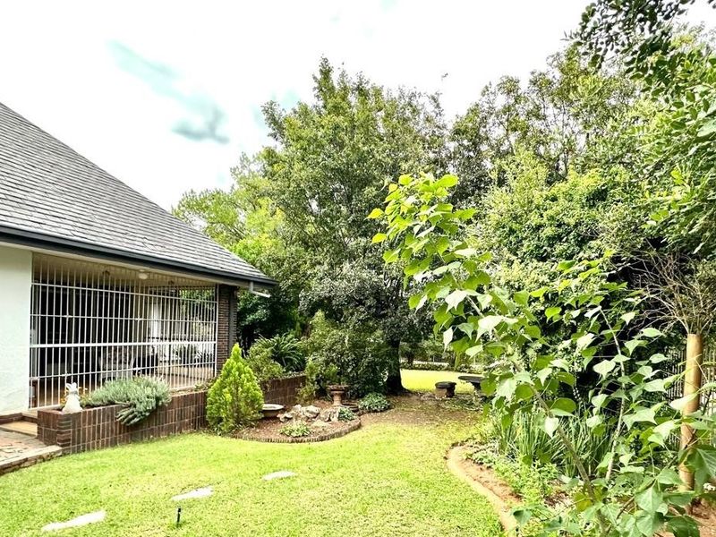 A view of the rear garden featuring a lawn, mature trees, a brick planter, and a covered patio area with security bars.