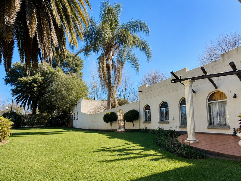 A well-maintained rear garden featuring a tiled patio, arched windows, and a pergola, set against a landscaped lawn and mature palm trees.