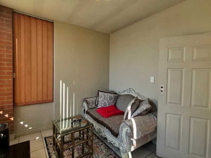 A living room featuring tiled flooring, vertical blinds, and an exposed brick accent wall, providing a functional space with potential for modernization.