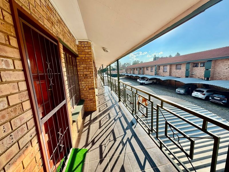 A covered exterior balcony featuring brick walls, tiled flooring, and a metal security balustrade overlooking the apartment complex parking area.