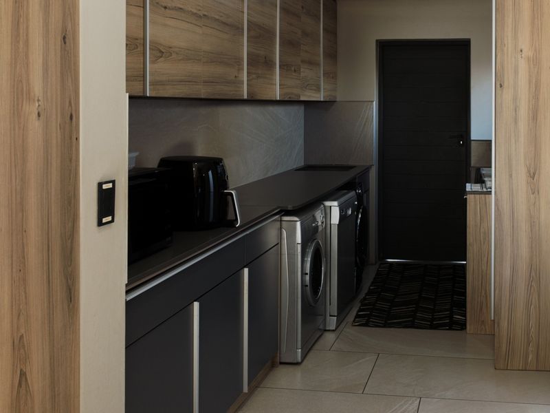 A modern, well-appointed scullery featuring sleek dark cabinetry, wood-grain accents, stone countertops, and dedicated space for laundry appliances.