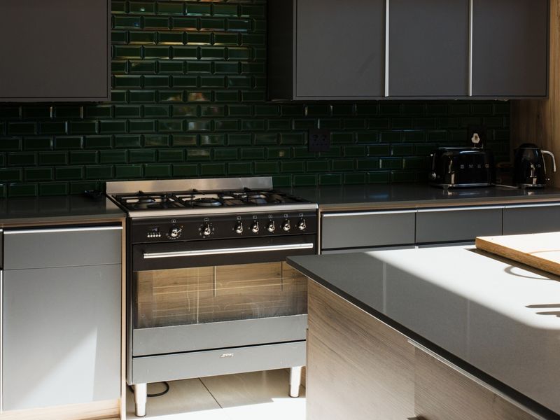 Modern kitchen featuring dark grey cabinetry, a striking green subway tile backsplash, stone countertops, and a high-end gas stove with oven.