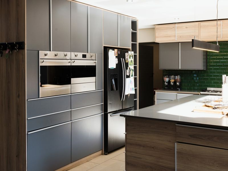 Modern kitchen featuring sleek grey cabinetry, wood-tone accents, a central island, and a striking green subway tile backsplash with integrated built-in appliances.
