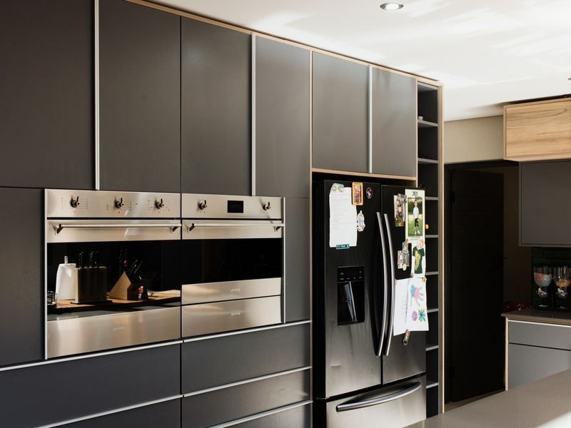 Modern kitchen featuring sleek dark cabinetry, built-in double ovens, stone countertops, and a stylish subway tile backsplash with integrated lighting.