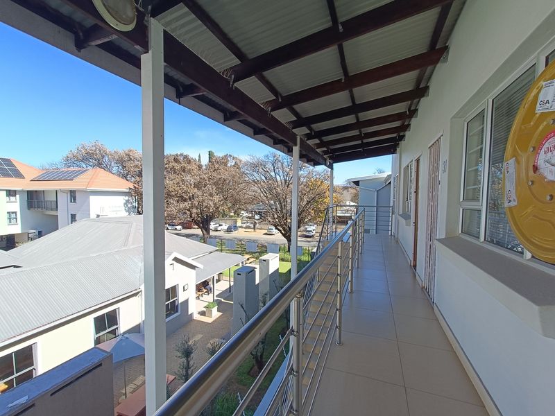 A covered balcony featuring tiled flooring, a stainless steel balustrade, and a corrugated metal roof, overlooking the apartment complex grounds.