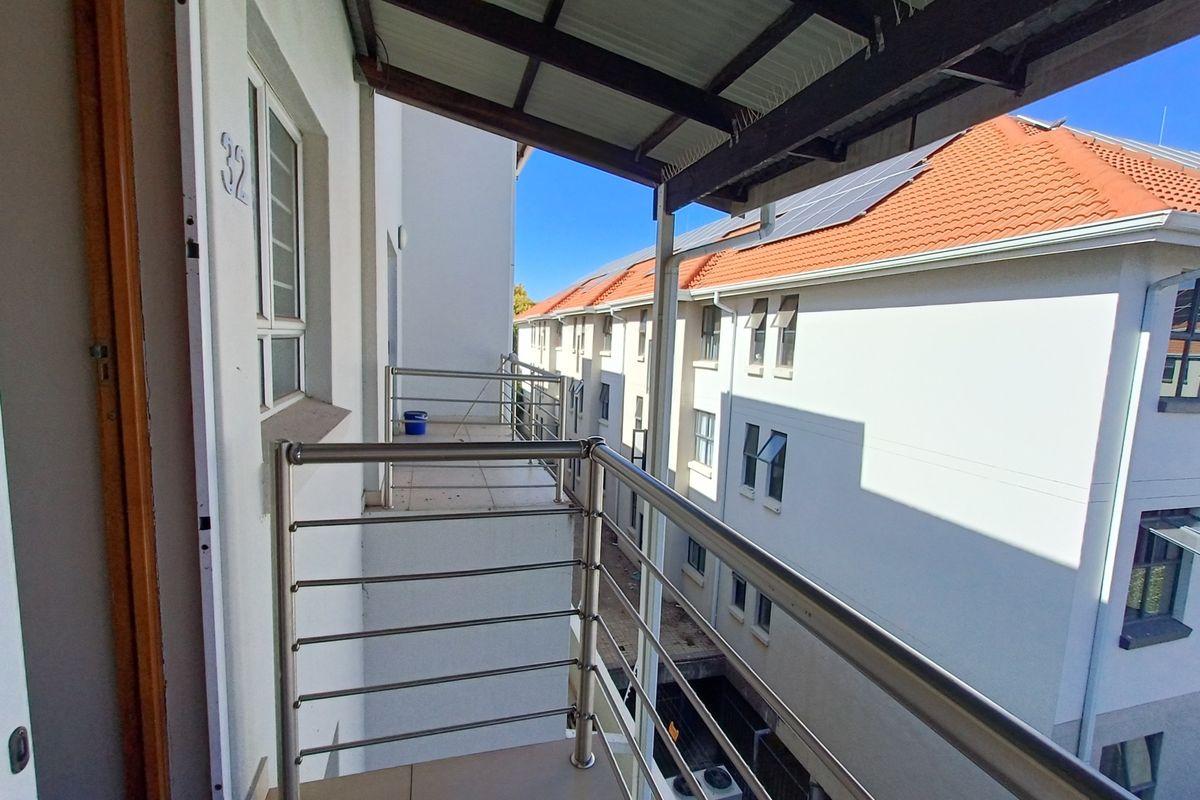 A covered balcony featuring tiled flooring and a stainless steel balustrade, overlooking the apartment complex courtyard.