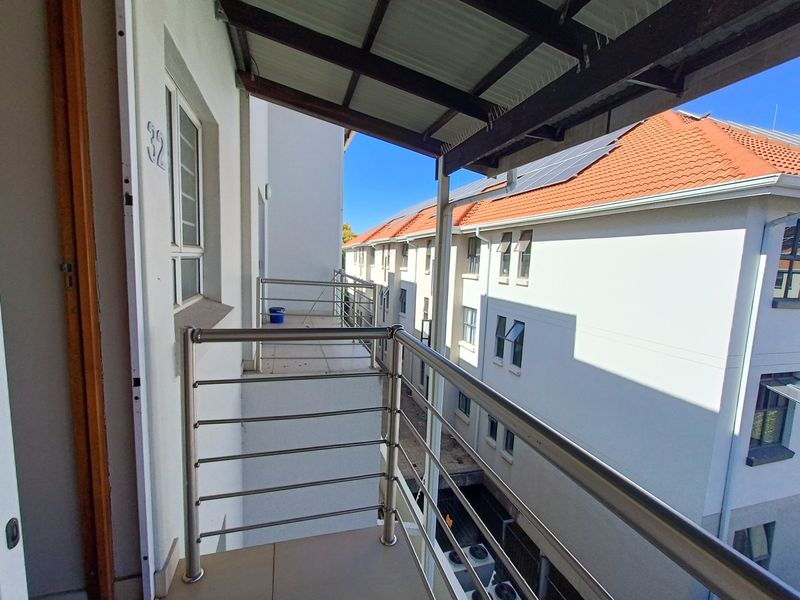 A covered balcony featuring tiled flooring and a stainless steel balustrade, overlooking the apartment complex courtyard.