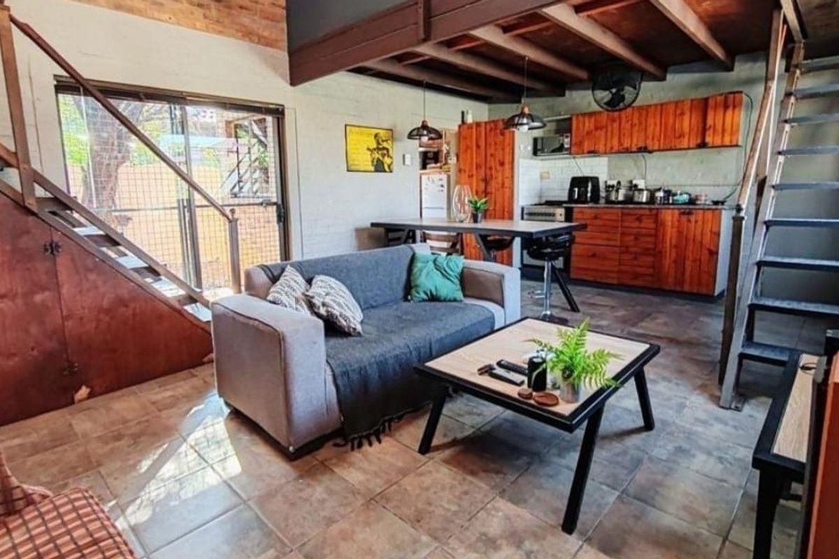 Open-plan living area featuring tiled floors, exposed timber ceiling beams, a metal staircase, and a functional kitchen with wooden cabinetry.