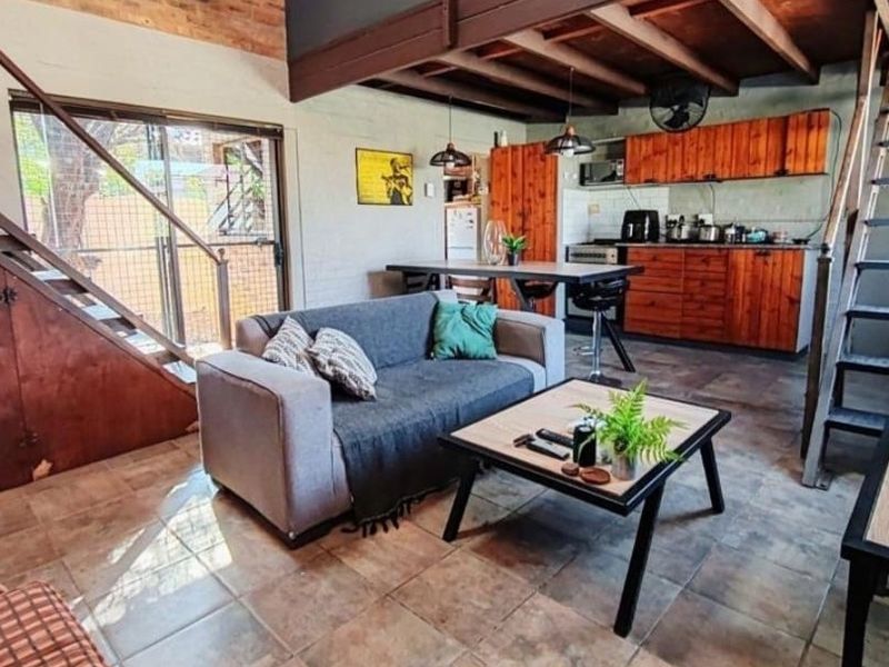 Open-plan living area featuring tiled floors, exposed timber ceiling beams, a metal staircase, and a functional kitchen with wooden cabinetry.