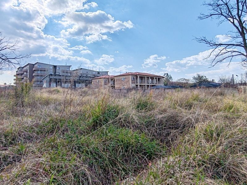 A view of a vacant residential plot featuring overgrown grass and a boundary wall, situated in a developed neighborhood.