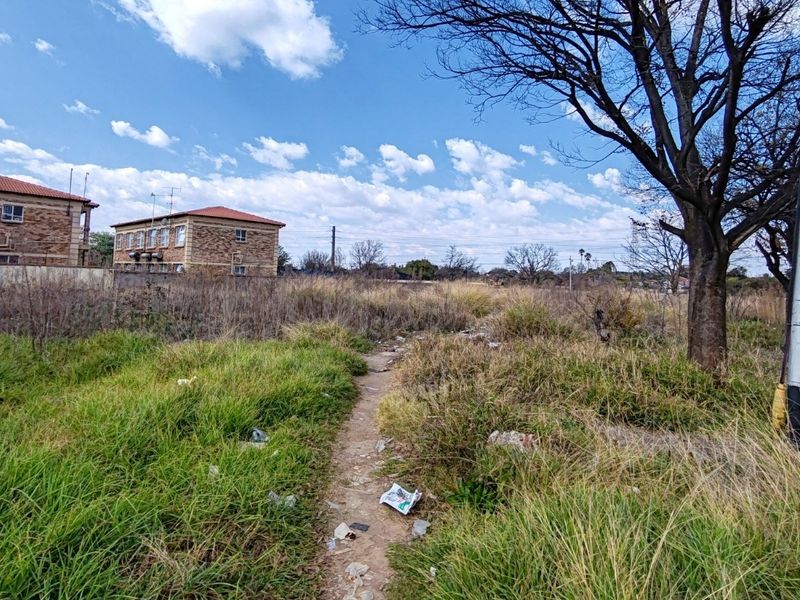 A view of a vacant residential plot featuring overgrown grass, a mature tree, and a utility pole, with neighboring residential buildings in the background.