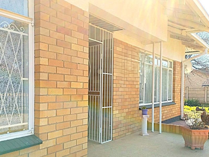 Front elevation of a brick house featuring a covered patio, security gate, and metal window frames, set within a residential garden.