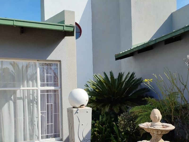 A rear exterior view of the property featuring plastered walls, green metal roofing, security window bars, and a decorative stone fountain in the garden.