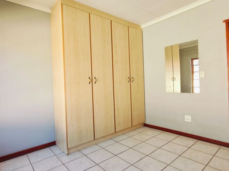 A clean, empty bedroom featuring light wood-finish built-in cupboards, neutral tiled flooring, and a wall-mounted mirror.