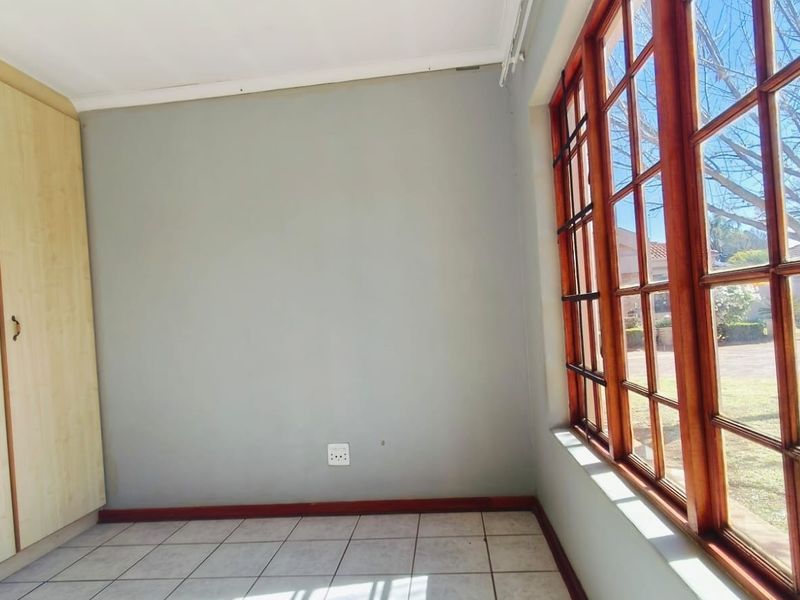 A bright, empty bedroom featuring tiled flooring, built-in cupboards, and large wooden-framed windows overlooking the garden.