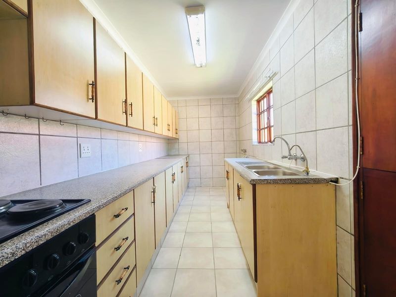 A functional galley-style kitchen featuring light wood cabinetry, laminate countertops, a double sink, and built-in cooking appliances with neutral tiled finishes.