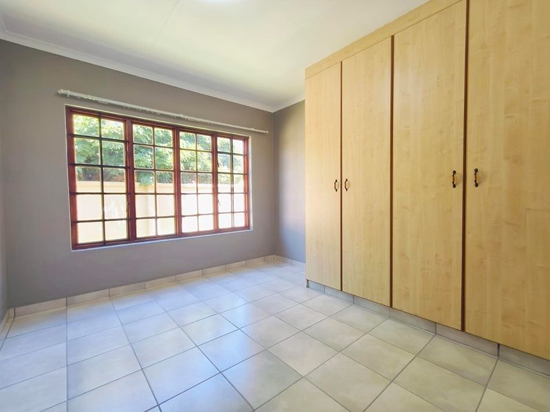 A bright, empty bedroom featuring neutral wall paint, tiled flooring, and extensive built-in wooden cupboards, offering a clean and versatile living space.