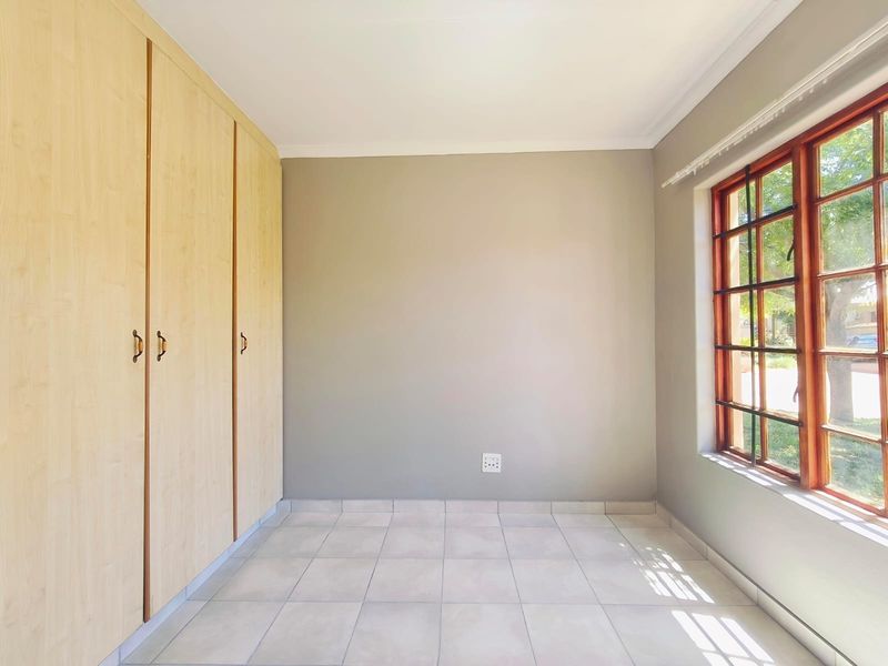 A bright, empty bedroom featuring neutral tiled flooring, built-in wooden cupboards, and a large window with a garden view.
