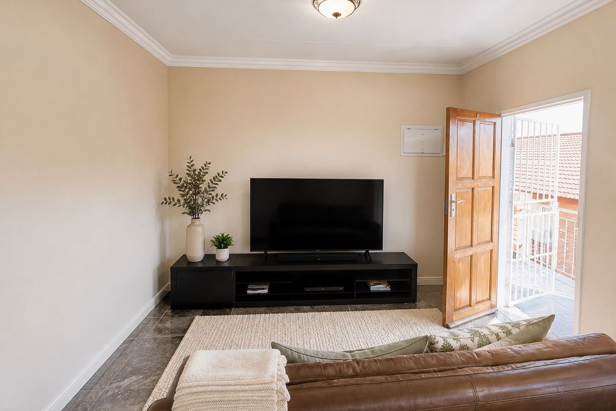 A well-maintained living room featuring tiled flooring, decorative ceiling cornicing, and direct access to an outdoor balcony through a timber door with security gate.