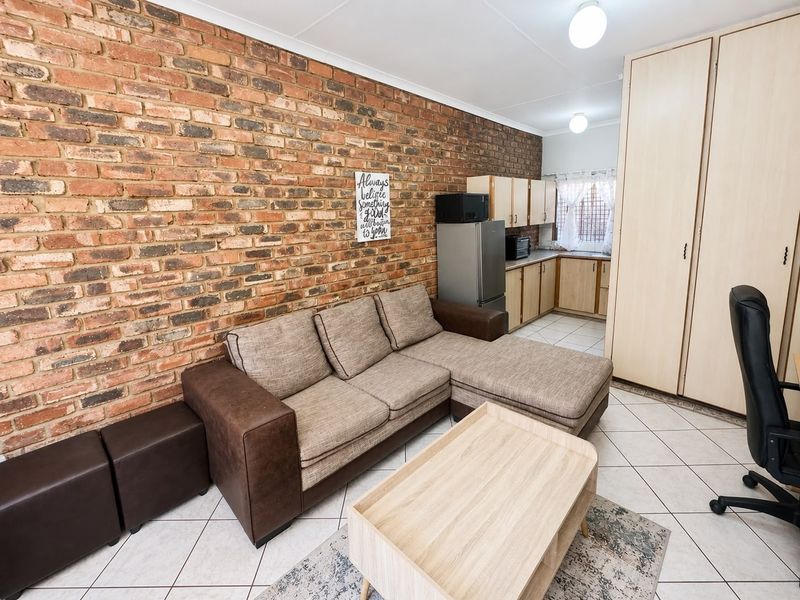 Open-plan living area featuring an exposed brick feature wall, tiled flooring, and a compact kitchen with built-in cabinetry and storage cupboards.